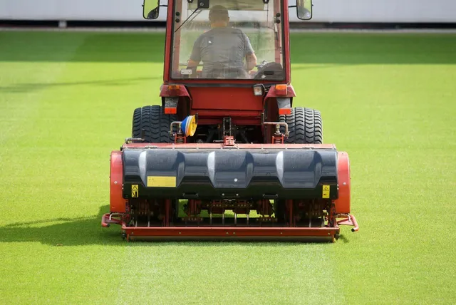 A red tractor-mounted lawn mower cuts a bright green field in a stadium.
